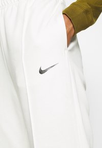 Bílé Nike tepláky mají texturovaný povrch s černým logem Nike na levém stehně a bočními kapsami s elastickým pasem.