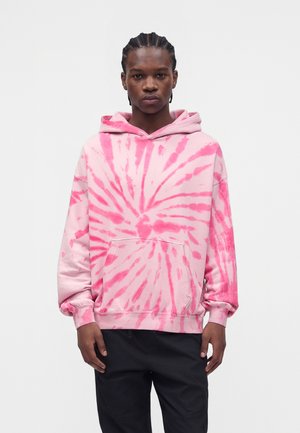 Jeune homme aux cheveux tressés portant un sweat à capuche tie-dye rose et rouge et un pantalon noir, debout devant un fond blanc uni.