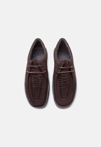 Chaussures en daim marron foncé avec détails tissés à l'avant et deux lacets en cuir horizontaux, présentées côte à côte sur un fond blanc.