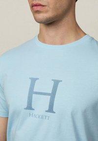 T-shirt en coton bleu clair avec un grand logo gris "H" et le mot "Hackett" en dessous, avec un col rond et des manches courtes.