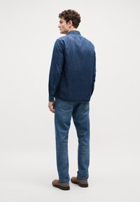 Tumša džinsa krekla ar pogām, valkāta virs gaišām džinsu biksēm. Apģērbs ir brīvā piegriezumā un vienkāršā dizainā. Izskatu papildina brūni apavi.