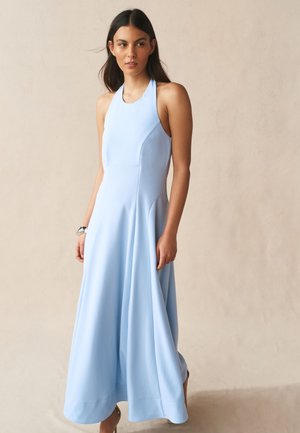 Nő, aki ujjatlan, világoskék halter nyakú maxiruhát visel és semleges bézs háttér előtt áll.