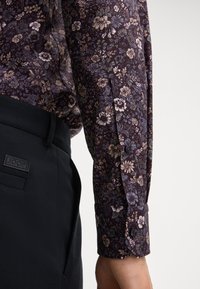 Langarmhemd mit einem dunklen lila Blumenmuster und hellen Akzenten, mit geknöpften Manschetten, kombiniert mit engen schwarzen Hosen, die das Logo präsentieren.