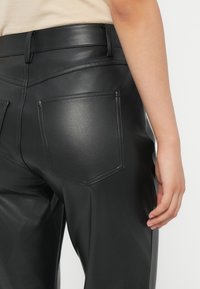 Detailní záběr na osobu v černých kalhotách z umělé kůže, kde je vidět zadní kapsa a část ruky, která odpočívá po boku.