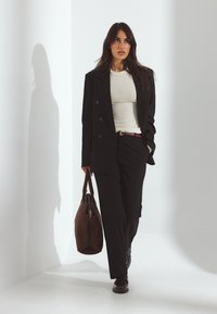 Frau in schwarzem Anzug und beigem Oberteil, die eine große braune Umhängetasche trägt, geht in einem weiß beleuchteten Raum in der Ecke entlang.