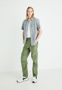Short-sleeve gray button-up shirt with two front pockets, paired with loose green cargo pants. Worn with white sneakers featuring black accents.