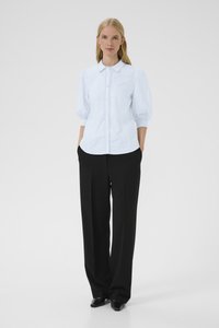 Donna in piedi contro uno sfondo bianco, che indossa una camicetta azzurra chiara con bottoni e maniche a sbuffo, e pantaloni neri a gamba larga, con le mani nelle tasche.