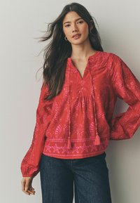 Blusa rossa con intricate fantasie rosa, maniche lunghe con polsini elastici, scollatura a V e tasselli decorativi sul davanti.