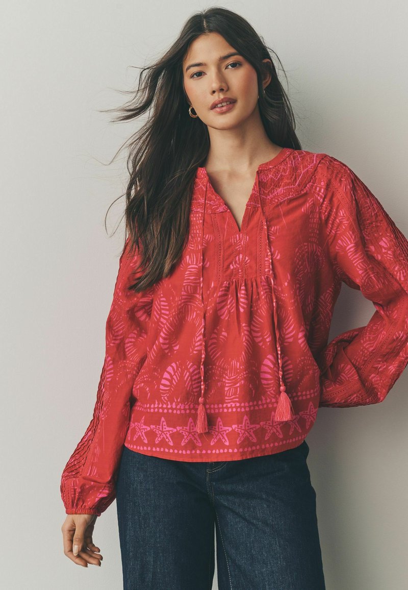 Blusa rossa con intricate fantasie rosa, maniche lunghe con polsini elastici, scollatura a V e tasselli decorativi sul davanti.