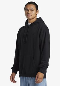Zwarte hoodie van zacht materiaal met een voorzak, een capuchon met trekkoord en een klein logo. Gecombineerd met lichtblauwe jeans.