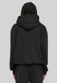 Sudadera negra con una gran capucha ajustable, hombros caídos y mangas con puños. Tejido suave y texturizado, corte relajado y diseño minimalista.