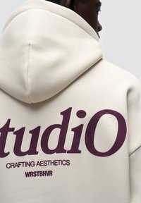 Felpa con cappuccio in morbido tessuto crema, con grande scritta color bordeaux sulla schiena: "studiO CRAFTING AESTHETICS WRSTBHVR."