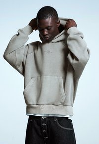 Világos bézs kapucnis pulóver elülső zsebbel, texturált anyagból és hímzett logóval. Párosítva sötét farmerral, melyen fémgombok találhatók.