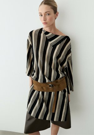 Donna con capelli biondi raccolti in uno chignon indossa una tunica a righe nere e beige, cintura marrone e gonna scura, in piedi contro una parete chiara e semplice.
