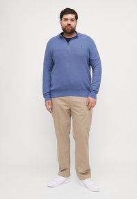 Lichtblauwe pullover met halve rits, geribbelde manchetten, gecombineerd met beige broek en witte sneakers, staand op een neutrale achtergrond.