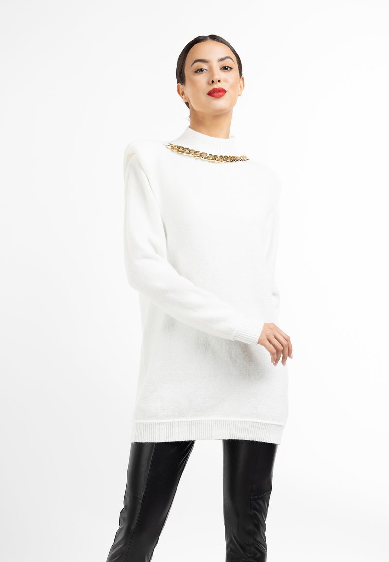 Witte oversized trui van zachte stof, met een hoge hals en gouden kettingdetail over de schouders. Gekozen in combinatie met zwarte leren broek.