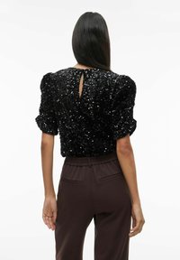 Melna blūze ar spīdīgām pērlītēm, pūkainām piedurknēm un atvērumu atpakaļ. Kombinēta ar brūnām biksēm ar elastīgu jostasvietu. Gluda tekstūra.