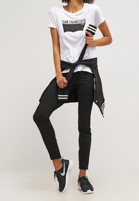 Persona vistiendo una camiseta blanca de "San Francisco", pantalones negros, una chaqueta negra atada a la cintura y zapatillas Nike negras con suelas blancas.
