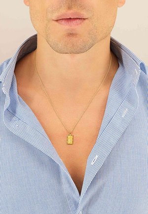 Collier en or avec un pendentif rectangulaire, gravure détaillée, sur une chaîne fine, porté sur une chemise à rayures bleues avec un col ouvert.