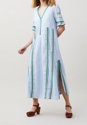 Vrouw draagt een lichtblauwe maxi-jurk met groene verticale borduur, pofmouwen, een zijsplit en bruine sandalen met hakken, staand tegen een effen achtergrond.
