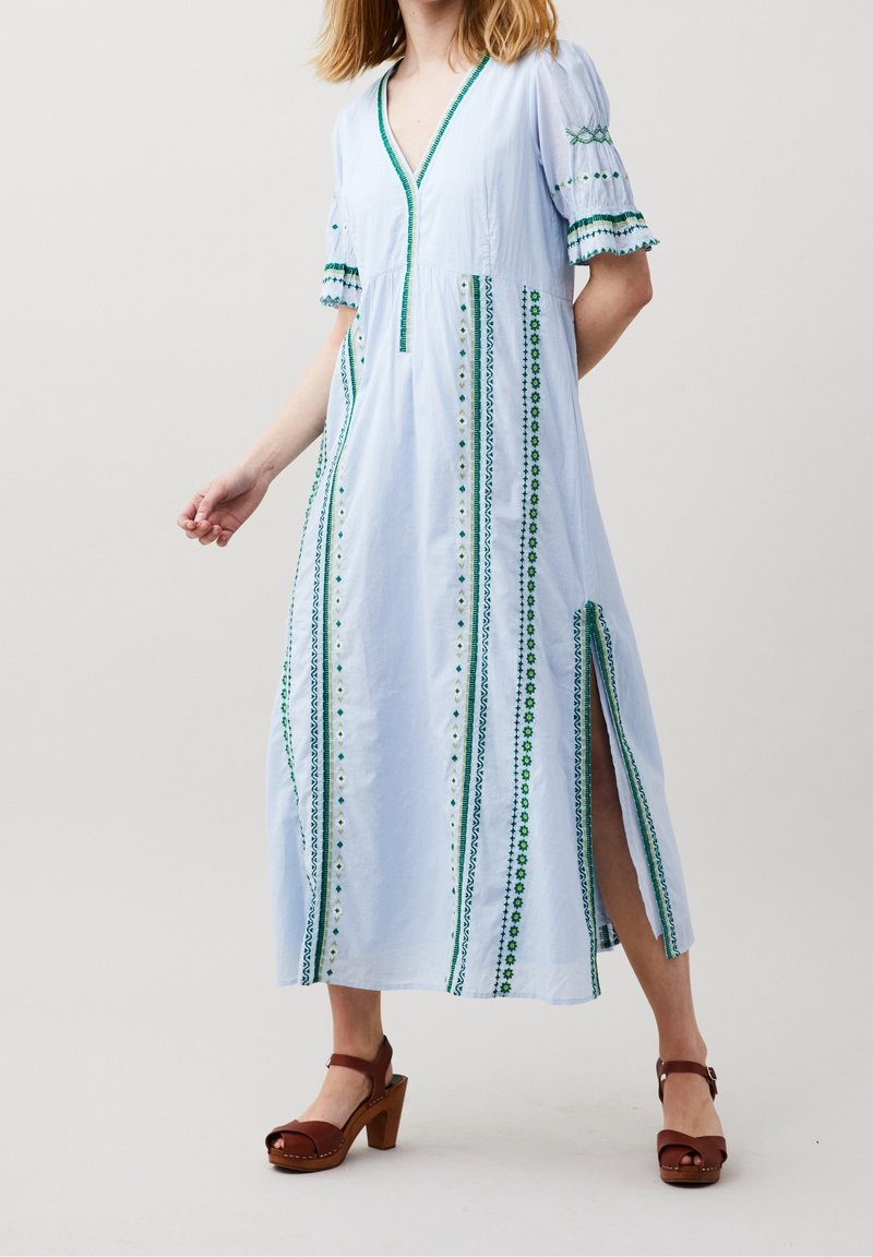 Femme portant une robe longue bleu clair avec broderie verticale verte, manches bouffantes, fente latérale et sandales marron à talons, debout devant un fond uni.