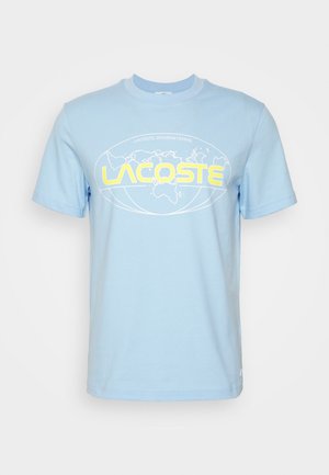 Lyseblå T-shirt med korte ærmer med gul LACOSTE tekst og et hvidt verdenskort design inde i en oval på brystet.