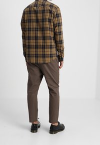 Brown and navy plaid long-sleeve shirt, buttoned cuffs, paired with brown slim-fit trousers and black shoes. Plain background.
