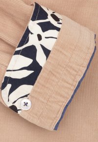 Beige corduroy stof met een bloemig patroon in het marineblauw aan de binnenste manchet. Een ronde witte knop met donker garen detail op de manchet.