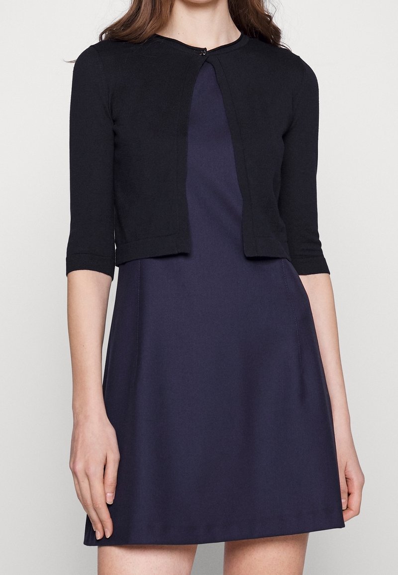 Femme portant une robe sans manches bleu marine avec un cardigan court noir ajusté aux manches longueur coude.