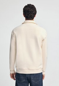 Felpa color crema con colletto classico, maniche lunghe, polsini a coste e orlo dritto, vista dal retro.