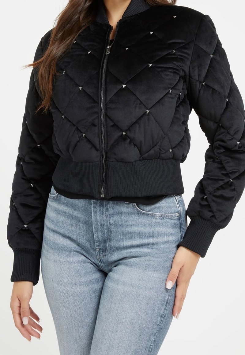 Femme portant une veste bomber matelassée noire avec de petits clous et un jean taille haute bleu clair, debout avec une main sur la hanche.