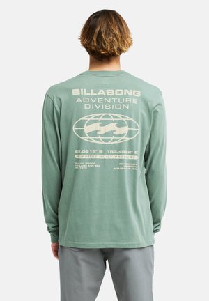 Persona con capelli castano chiaro indossa una maglietta verde a maniche lunghe con il testo "Billabong Adventure Division" e il logo del globo sulla schiena.