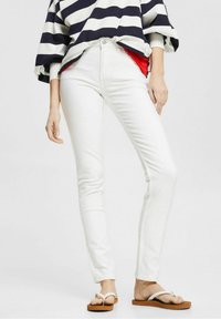 Bílé skinny džíny s vysokým pasem, kombinované s modro-bílým pruhovaným volným topem. Modelka má na sobě hnědé žabky. Hladká textura.