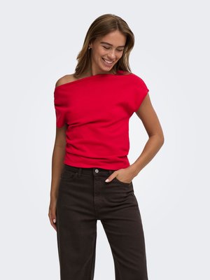 Kvinde smilende, iført en rød off-shoulder top og sorte bukser, med den ene hånd i lommen mod en ensartet lys baggrund.