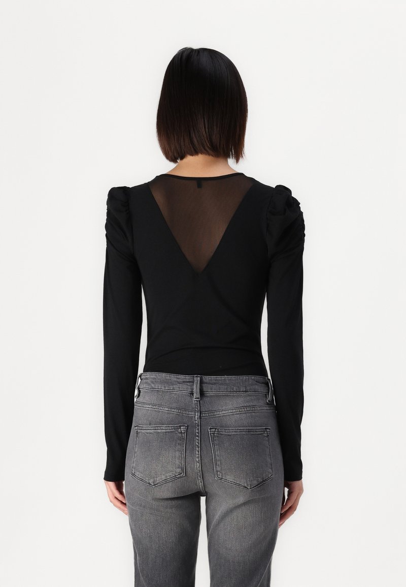 Schwarzes Langarmshirt mit einem V-förmigen, transparenten Rücken und puffierten Schultern; kombiniert mit grauen Jeans aus Denim mit Gesäßtaschen und einer figurschmeichelnden Passform.