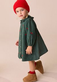 Groene corduroy jurk met kersenborduursel, knopen aan de voorkant en gerimpelde kraag. Gecombineerd met een rode gebreide beanie en tan enkellaarzen.