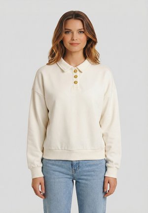 Mujer con sudadera tipo polo de manga larga color crema con tres botones dorados en forma de sol y jeans azul claro, de pie contra un fondo gris.