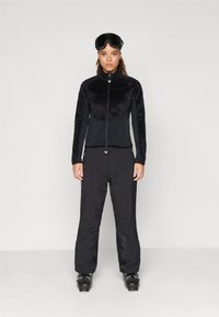 Must maapjakotiga fliiskardigan sujuva alumise osaga, koos mustade suusipükstega. Modell kannab suusaglasse, demonstreerides stiilset ja funktsionaalset disaini.