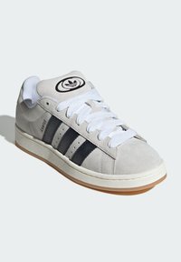 Sivi antilopasto obuća s bijelim vezicama, tri crne pruge i gumeni potplat. Ima okrugli logo na jeziku i teksturirane akcentne detalje.