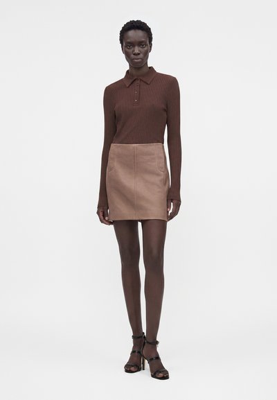 Γυναίκα που φοράει καφέ ριγωτό μακρυμάνικο πόλο, μπεζ μίνι φούστα με τσέπες και μαύρα σανδάλια με τακούνι, στέκεται σε απλό φόντο.