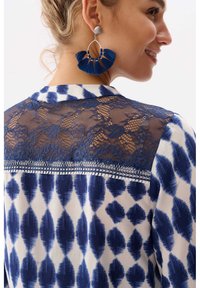 Blauw en wit gemusterd shirt met kant achter, kraag en decoratieve stiksels, gecombineerd met grote blauwe franjes oorbellen.