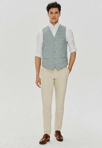 Jasnoniebieska kamizelka w kratę noszona na białej koszuli, beżowych chinosach i brązowych skórzanych butach. Kamizelka ma dekolt w serek i guziki.