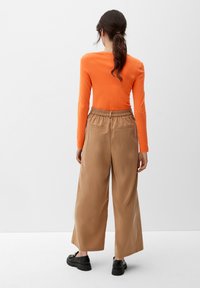 Maglietta arancione a maniche lunghe abbinata a pantaloni larghi beige con cintura elastica e tasche laterali; il modello indossa calzature nere.