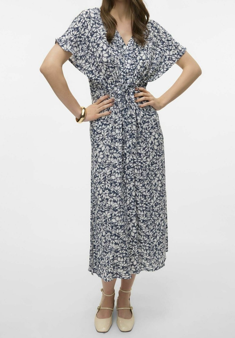 Květinové maxi šaty v bílé a námořnické modři, s krátkými rukávy, užším pasem a volnou siluetou. Nosené s béžovými sandály na popruhy.