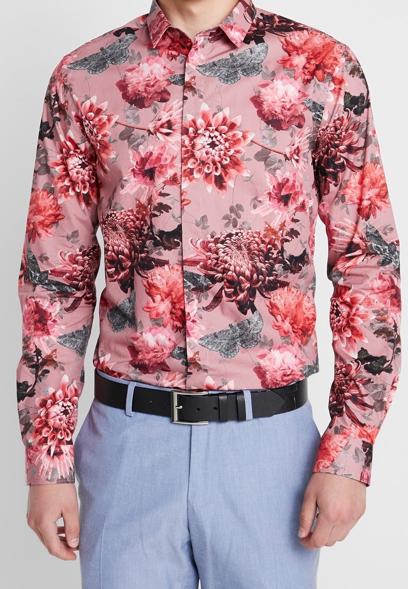 Bloemenprint overhemd met lange mouwen in tinten roze en rood met zwarte accenten. Heeft levendige bloemenpatronen en motdetails. Zwarte riem.