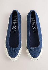 Paire de ballerines en denim bleu à enfiler avec bouts en caoutchouc blanc et petits nœuds à l'avant, étiquetées avec "NEXT" sur les semelles intérieures.
