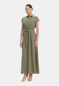 Abito maxi verde oliva con maniche corte, bottone anteriore, due tasche sul petto, cintura in vita e spacco laterale. Tessuto morbido con un design fluido.