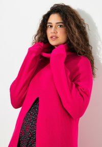 Pull en tricot fuchsia avec un col haut et des manches longues, présentant un design découpé à l'avant. Texture douce et coupe ample.