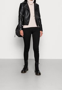 Giacca da biker in pelle nera sopra un dolcevita beige, abbinata a jeans skinny neri e stivali neri con lacci. Accessorio: borsa nera con texture.