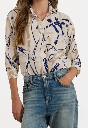 Femme portant une chemise à motifs beige et bleu marine boutonnée, rentrée dans un jean bleu délavé, avec une main dans la poche.